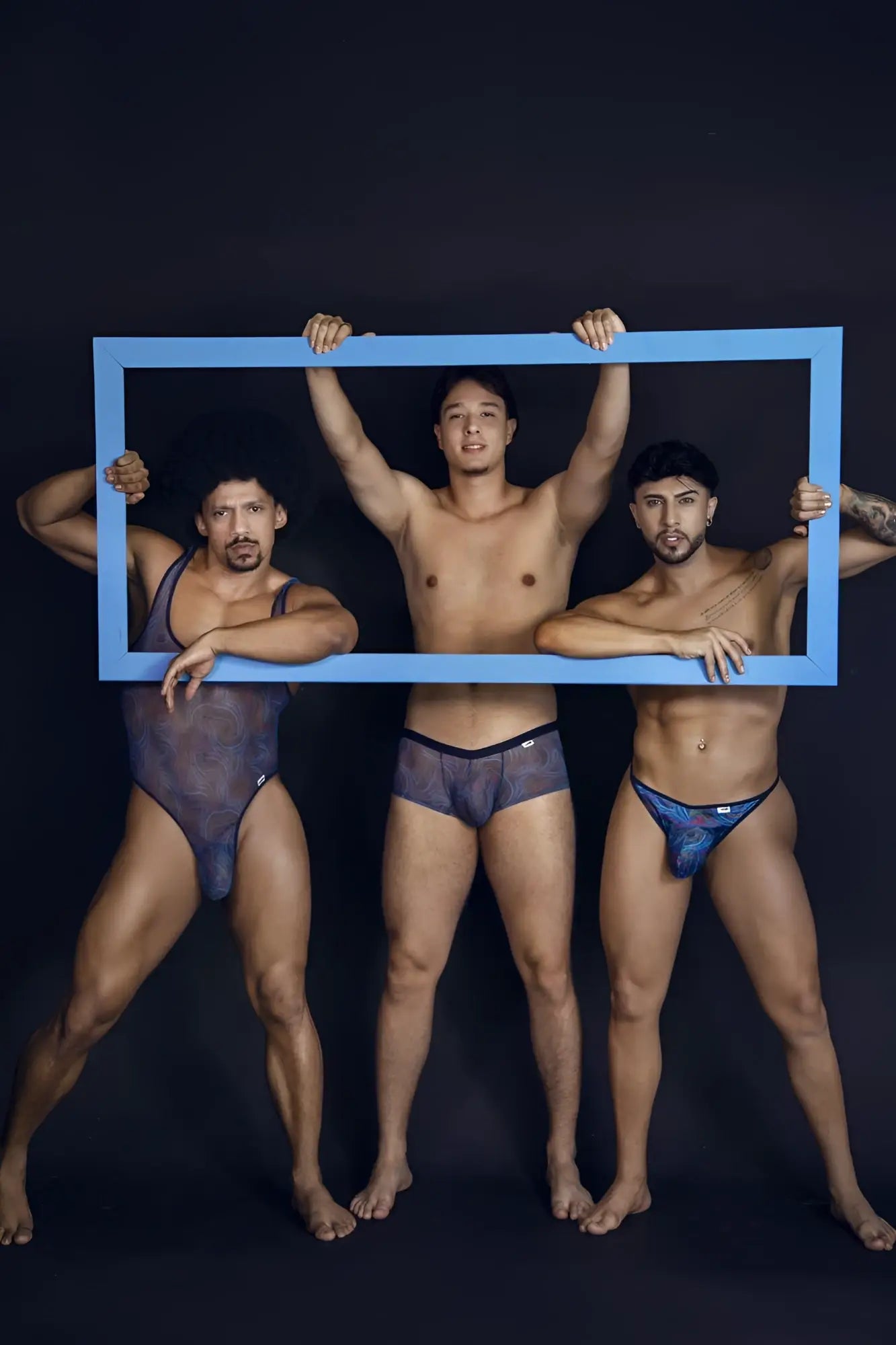 Three men holding a blue rectangular frame featuring candyMan 99875 seawater multicolor print thongs