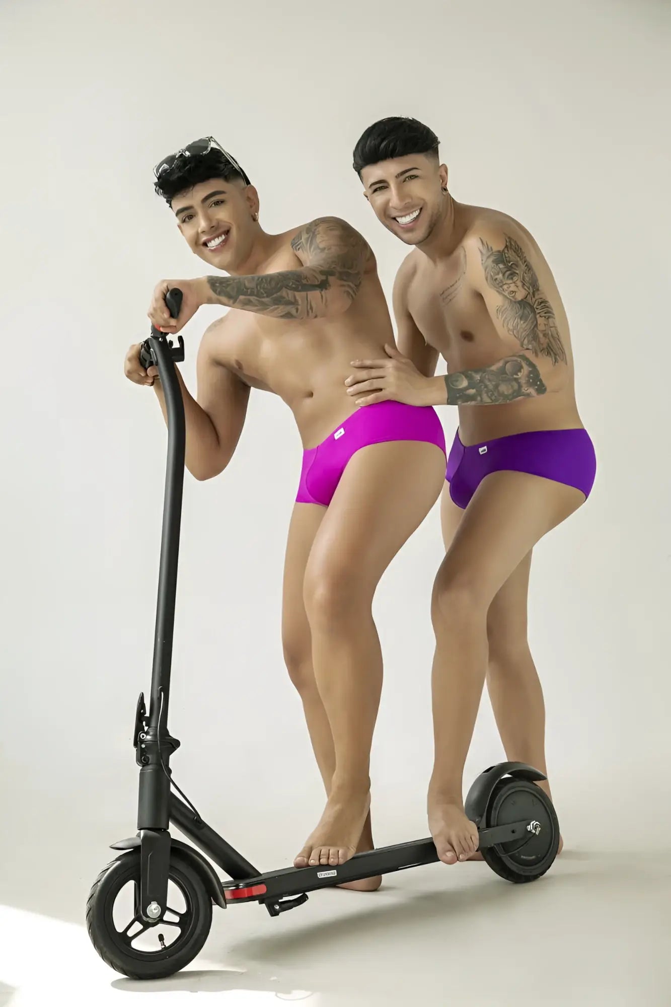 Two men with tattoos posing on a scooter wearing CandyMan magenta trunks in a form-fitting, low-rise style.