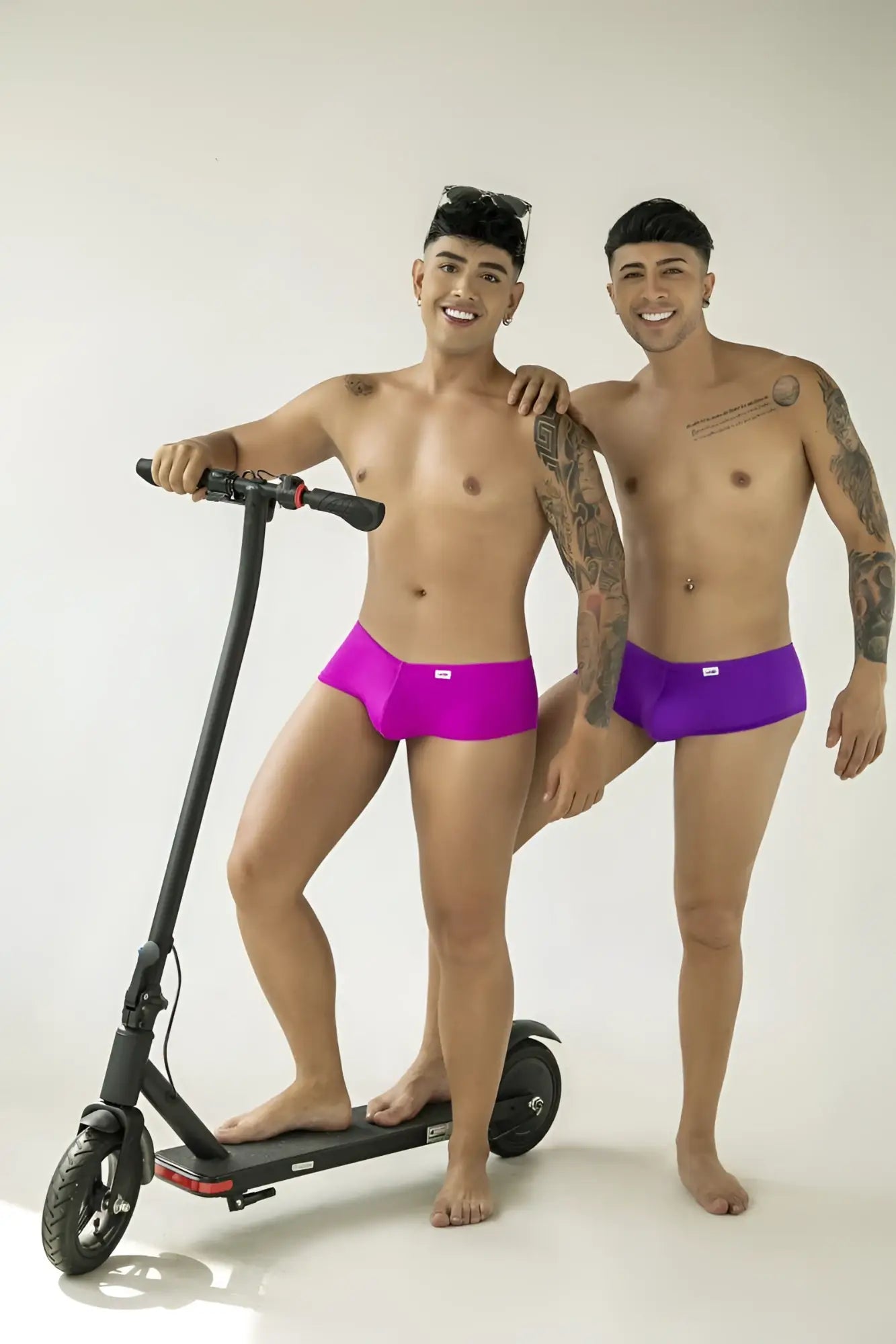 Two men wearing CandyMan magenta trunks pose on a white background, one with a scooter, showing a form-fitting style.