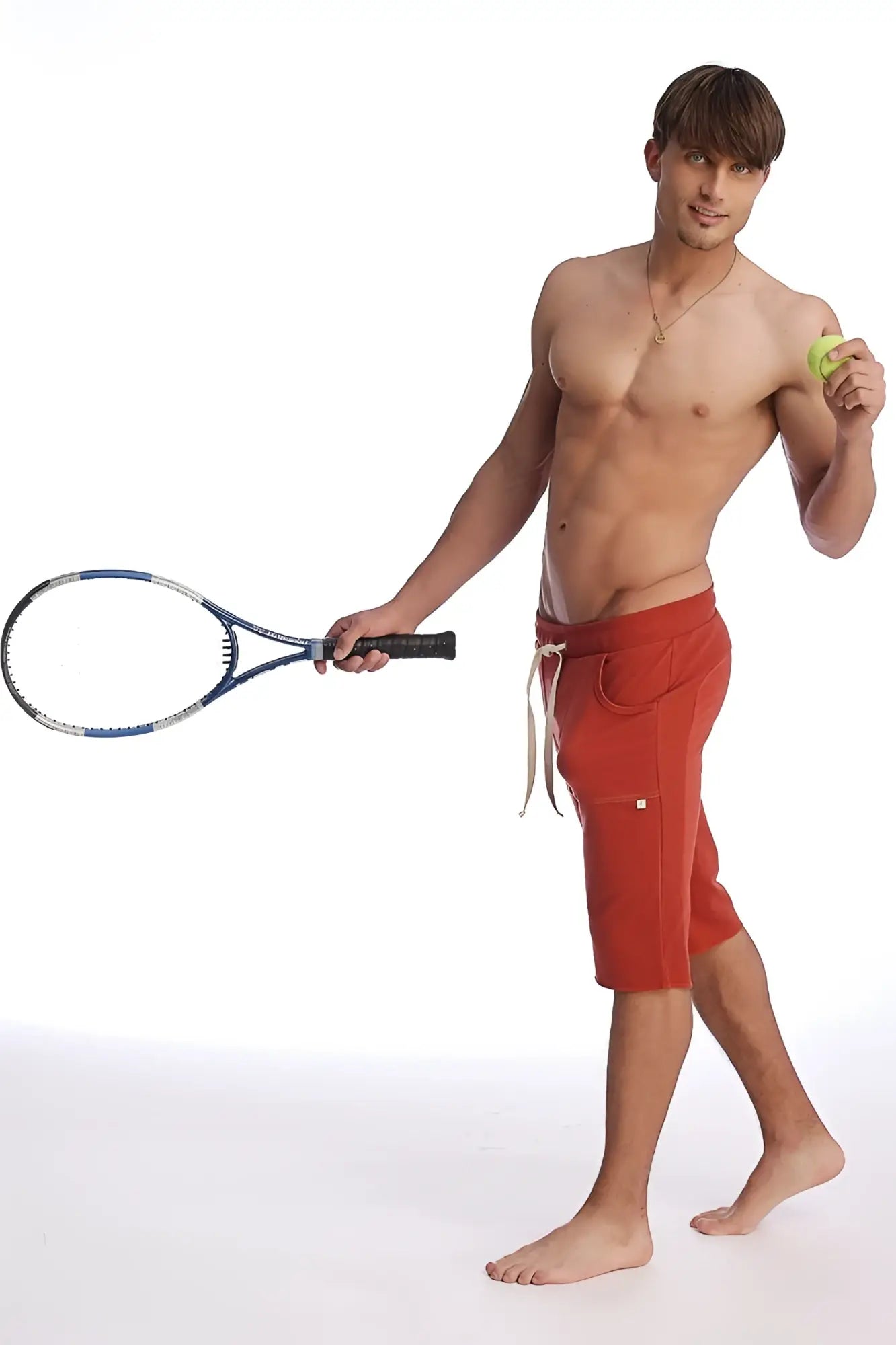 A man wearing cinnabar eco-track shorts with white drawstrings holding a blue tennis racket and yellow tennis ball