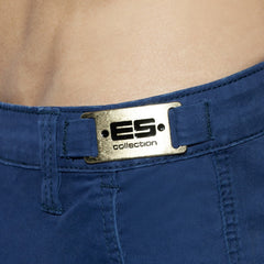 Close-up of blue cargo shorts with a gold metallic ES Collection logo plaque on the waistband.
