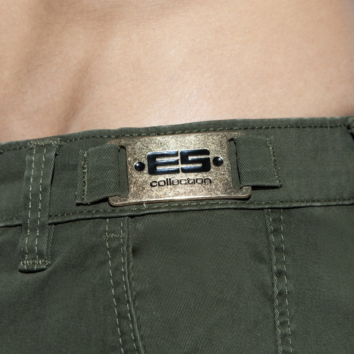 Close-up of olive green ES Collection cargo shorts with a metal brand plaque on the waistband.