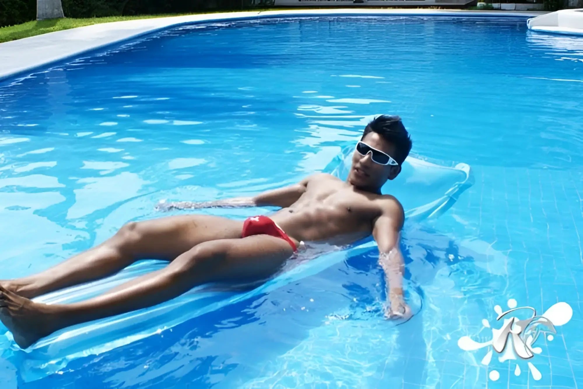 A man wearing red icker sea lifeguard micro slip bikini swimwear floats on a blue inflatable pool float in clear water