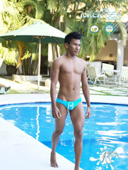 A man stands by a pool wearing icker sea lifeguard slip bikini swimwear in bright turquoise with white drawstrings