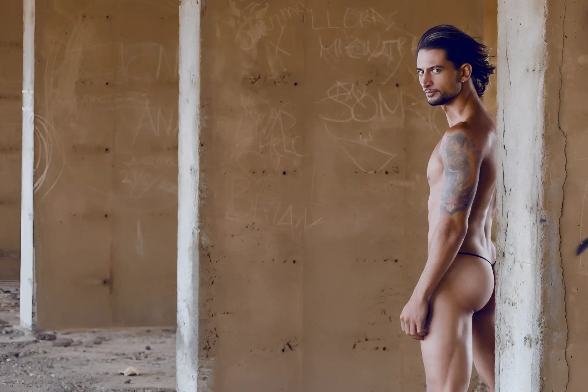 A muscular man with long dark hair and tattoos stands nude against graffiti wearing justin simon xsj02 classic turquoise g-string bulge