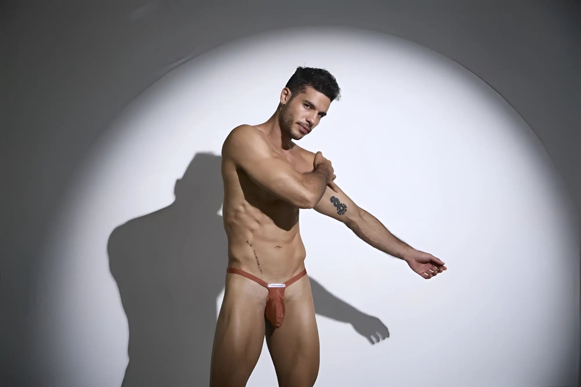 A man wearing a reddish-brown thin-strapped jockstrap with a small white label front in JUSTIN SIMON XSJBU02 ochre bulge thong