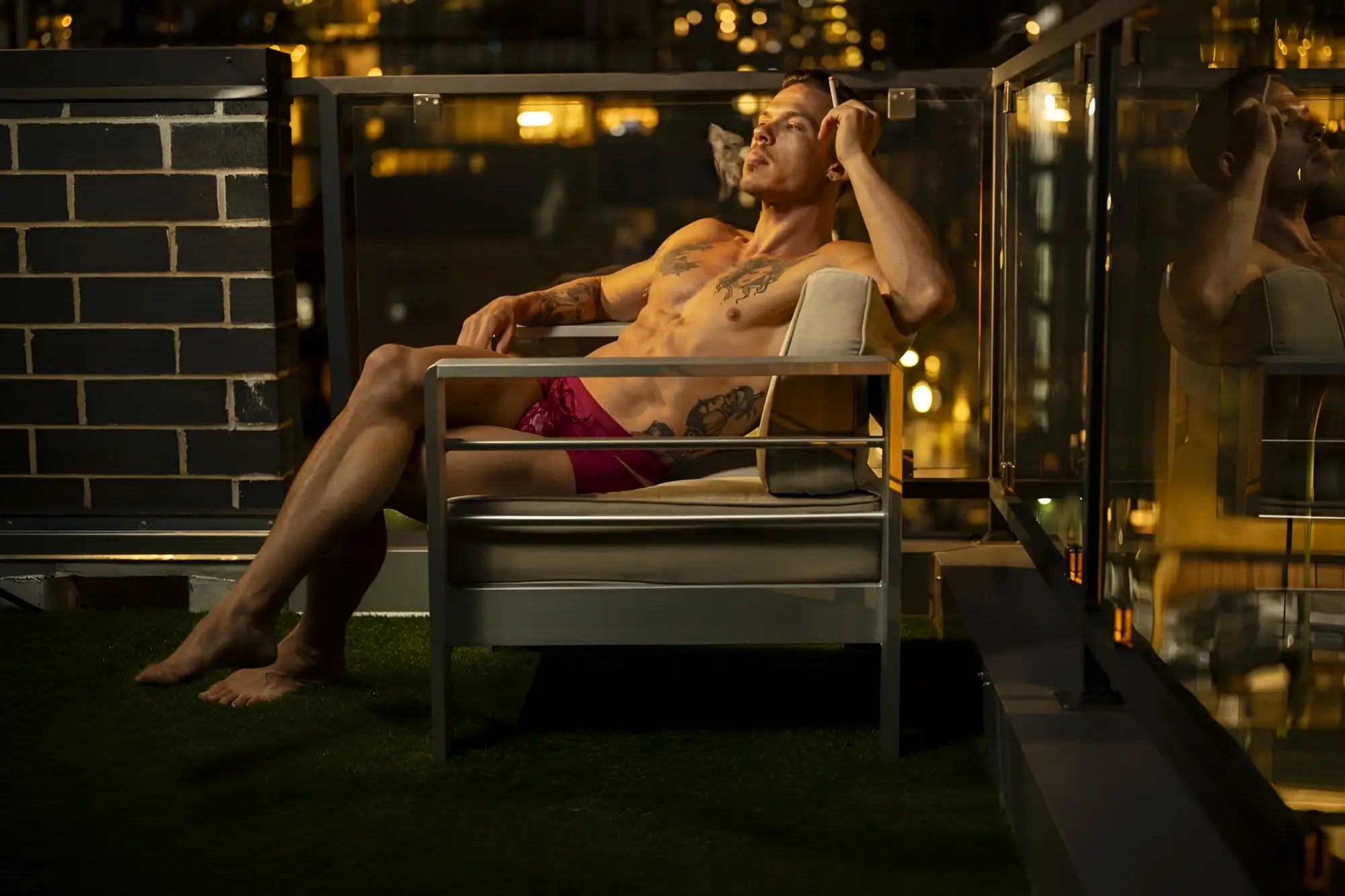 A man with tattoos lounges in a modern metal chair wearing male power 126-289 lucifer cut out shorts in burgundy