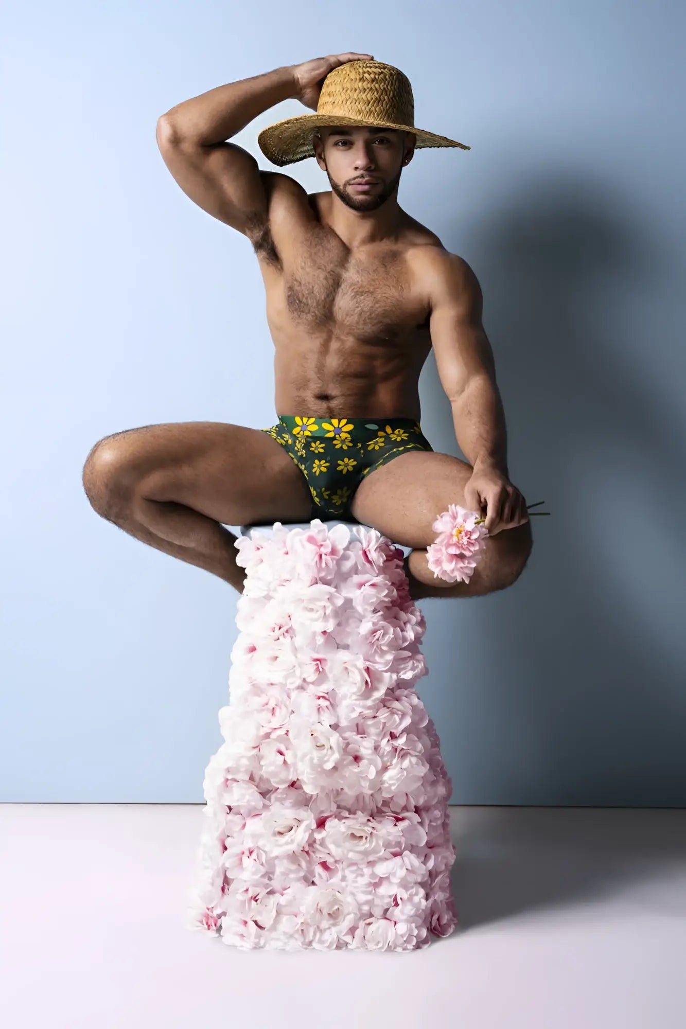 A man wearing green petal power pouch shorts with yellow daisy print sits on a pedestal of pink roses