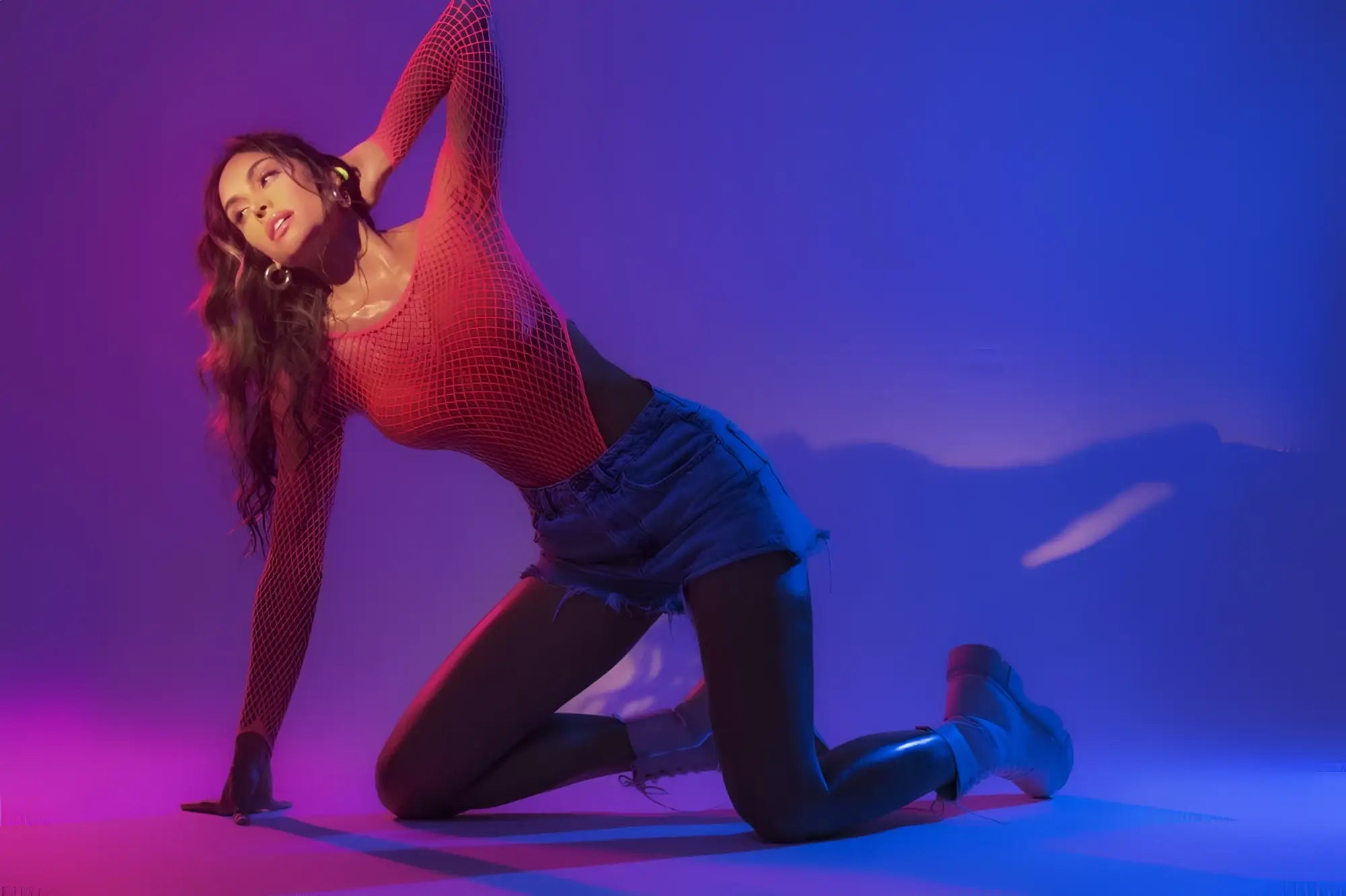 A woman poses in a neon pink mapale 1119 long sleeve fishnet bodysuit with red mesh and denim shorts under purple lighting