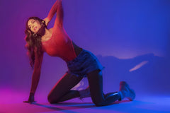 A woman poses in a neon pink mapale 1119 long sleeve fishnet bodysuit with red mesh and denim shorts under purple lighting