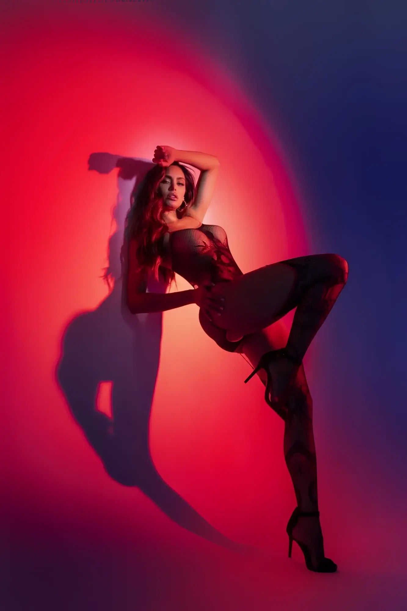 A woman wearing a black red Mapale 1154 snake fishnet bodysuit with lace details and heels against a red blue backdrop