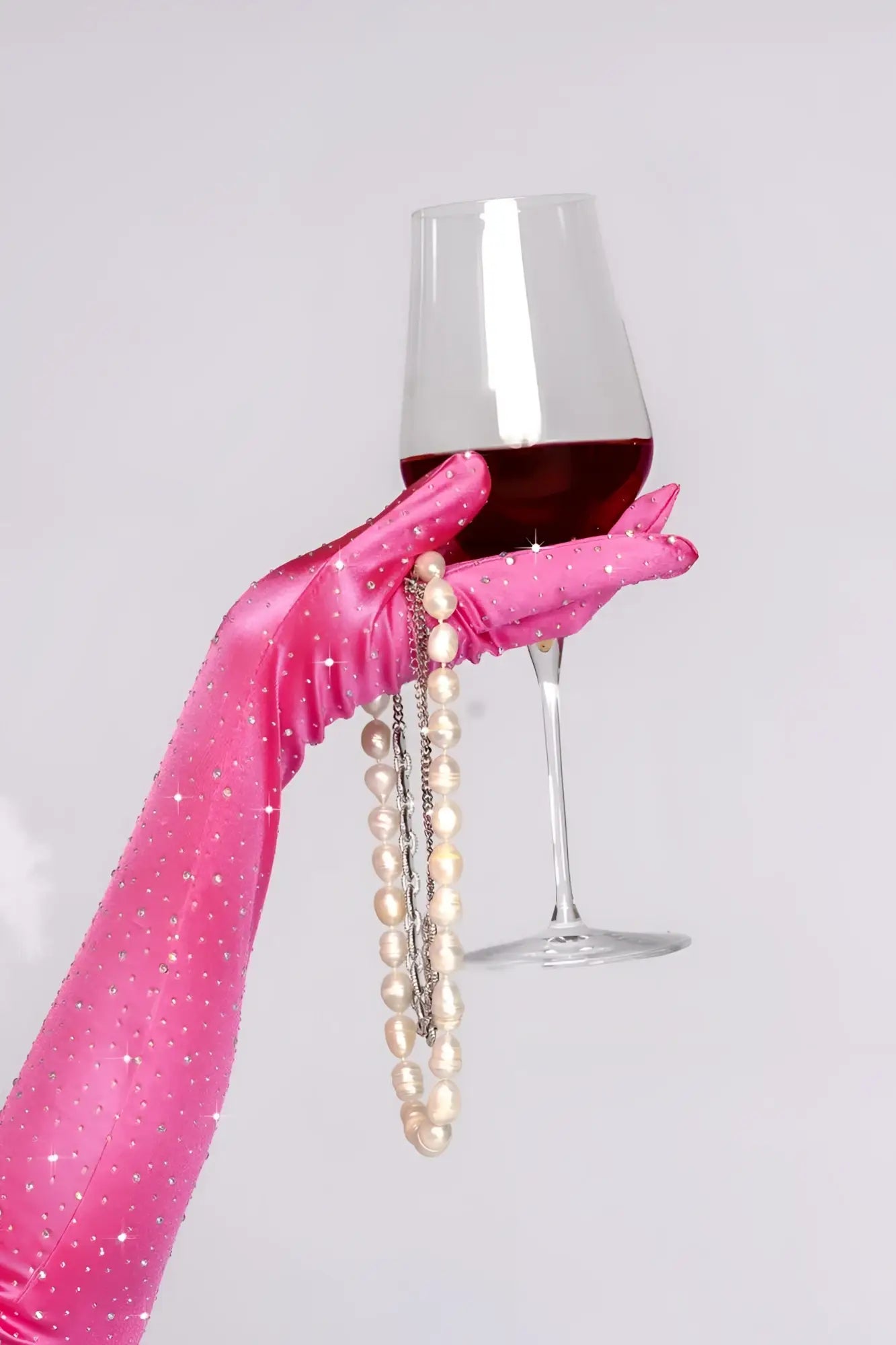 A hand wearing mapale pink satin rhinestone opera glove holds a red wine glass with pearl necklace accents