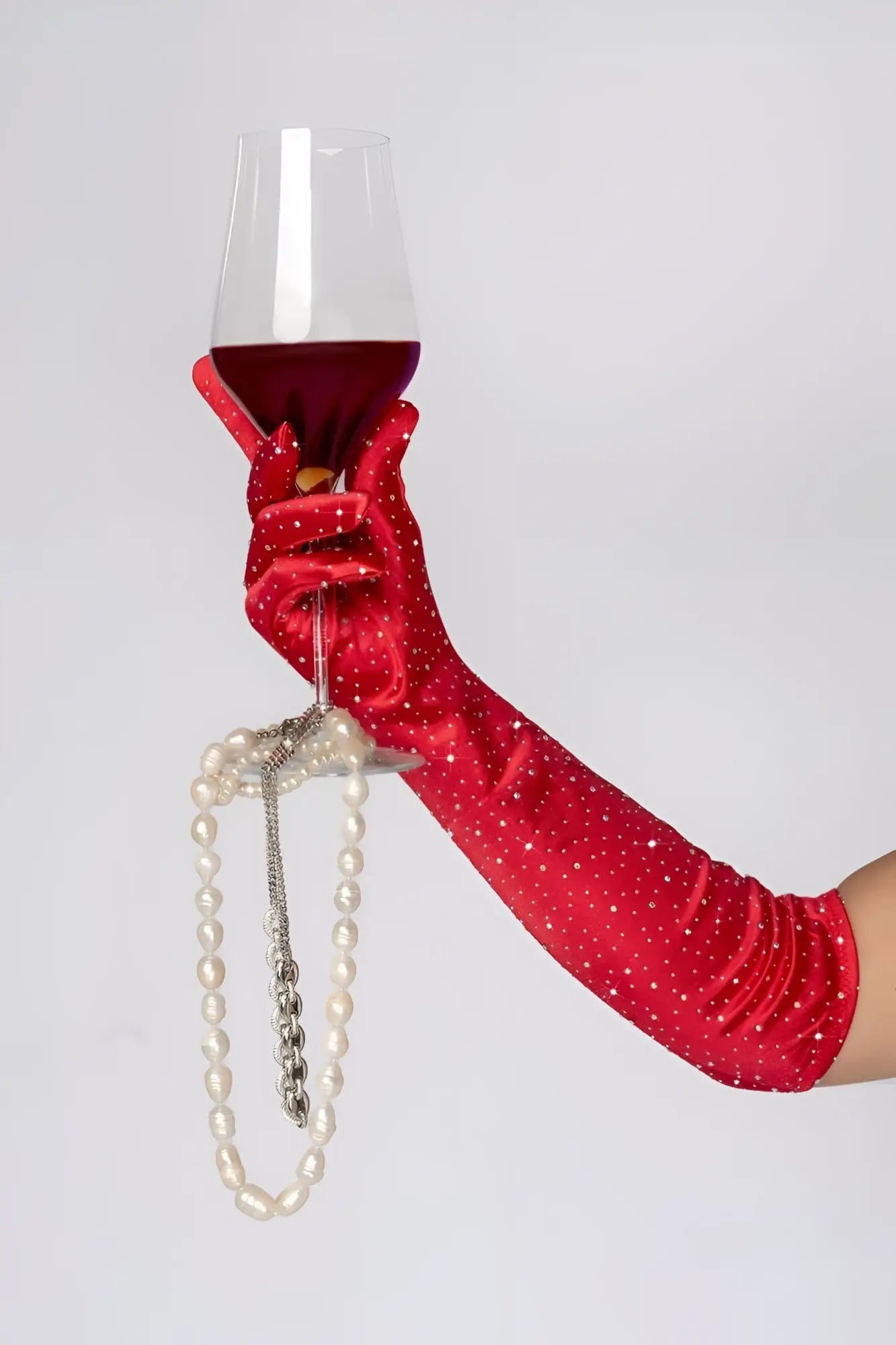 A hand in a red mapale 135 satin rhinestone opera glove holding a wine glass with a dangling pearl necklace