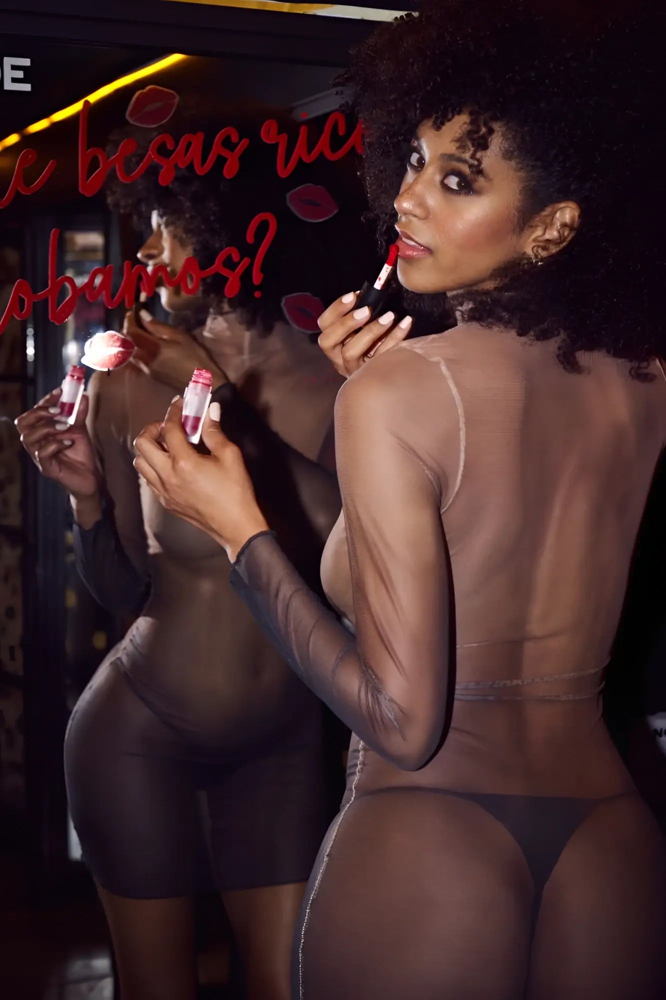A woman with voluminous curly hair wears a sheer black Mapale 40000 bodysuit in midnight shadow holding red lipstick
