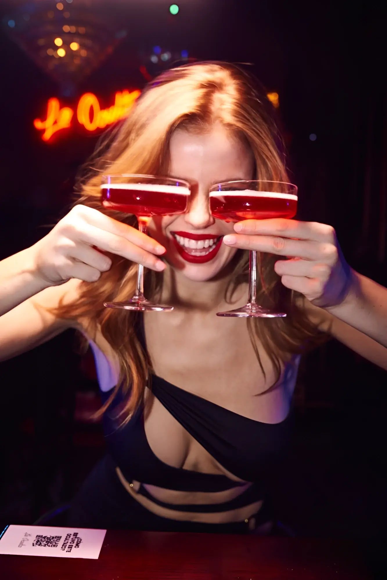 A woman with blonde hair holds two red drink-filled coupe glasses wearing a black mapale 40006 dress