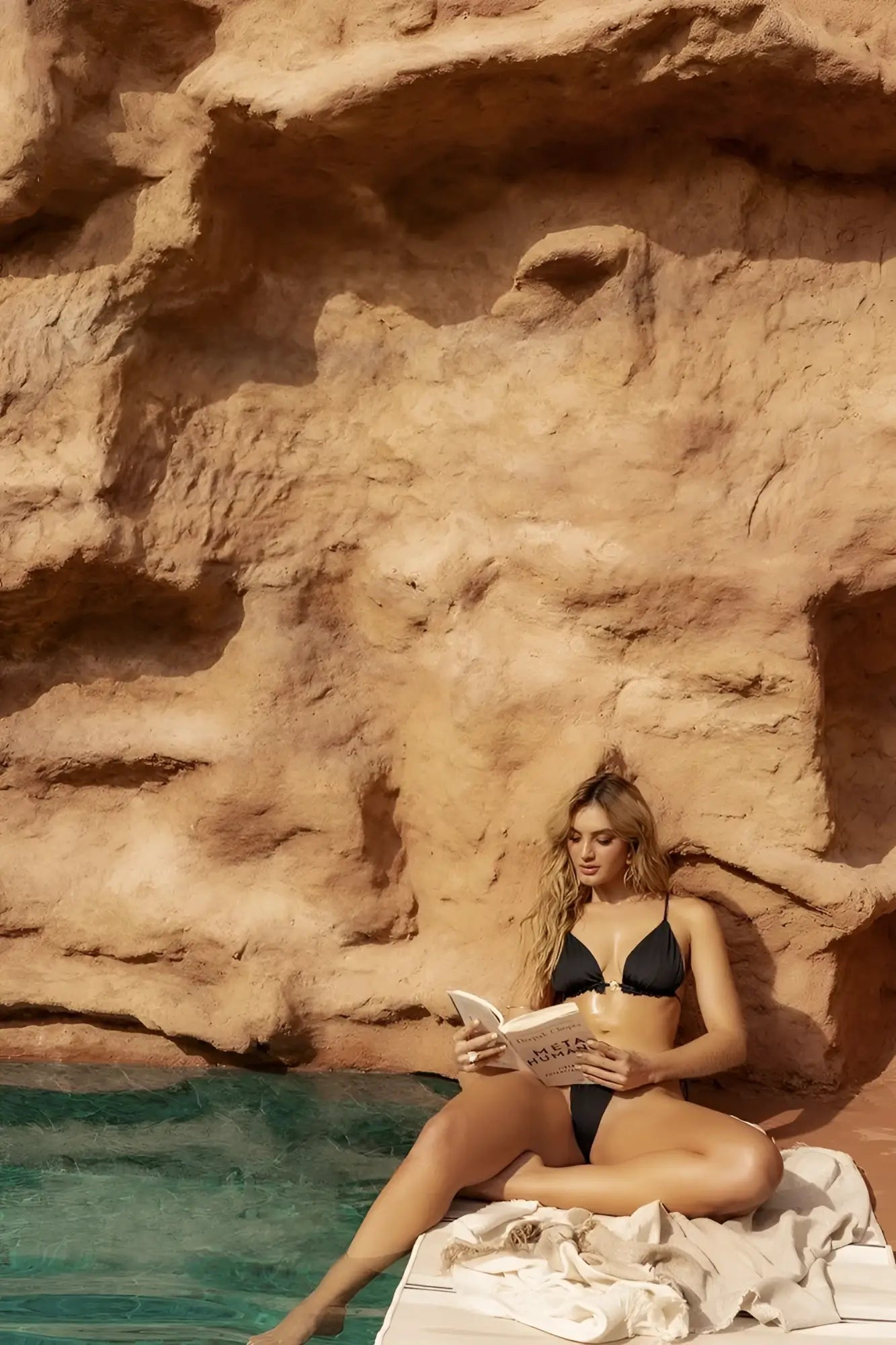 A woman sits poolside wearing a black Mapale 67166 two piece swimsuit reading a book with milia on the cover