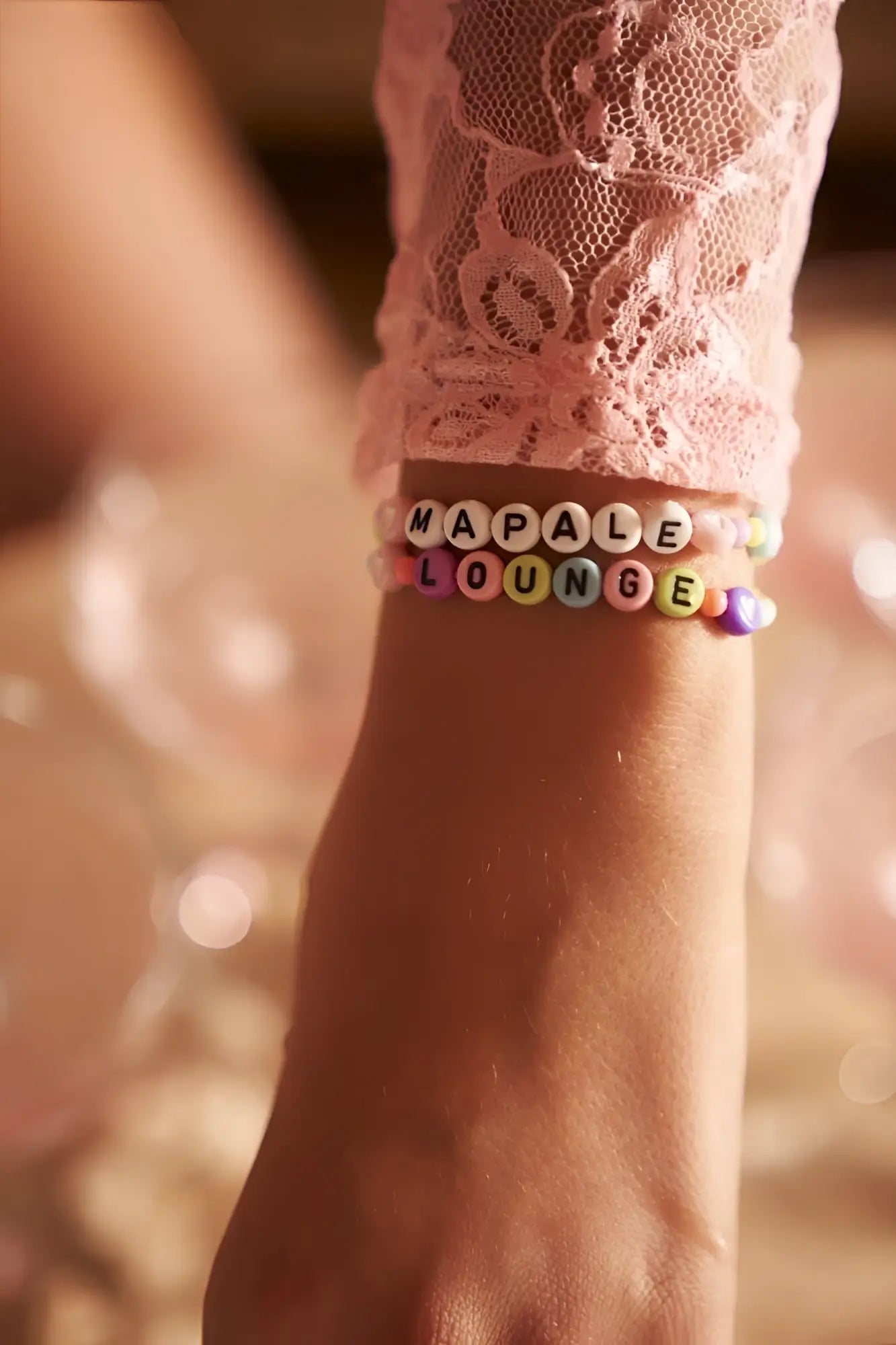 A colorful beaded bracelet with alphabet beads spelling mapale lounge worn on a wrist with a rose color sleep romper