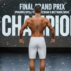 A muscular man wearing gray metroMuscleWear insignia physique board shorts stands with his back to the camera