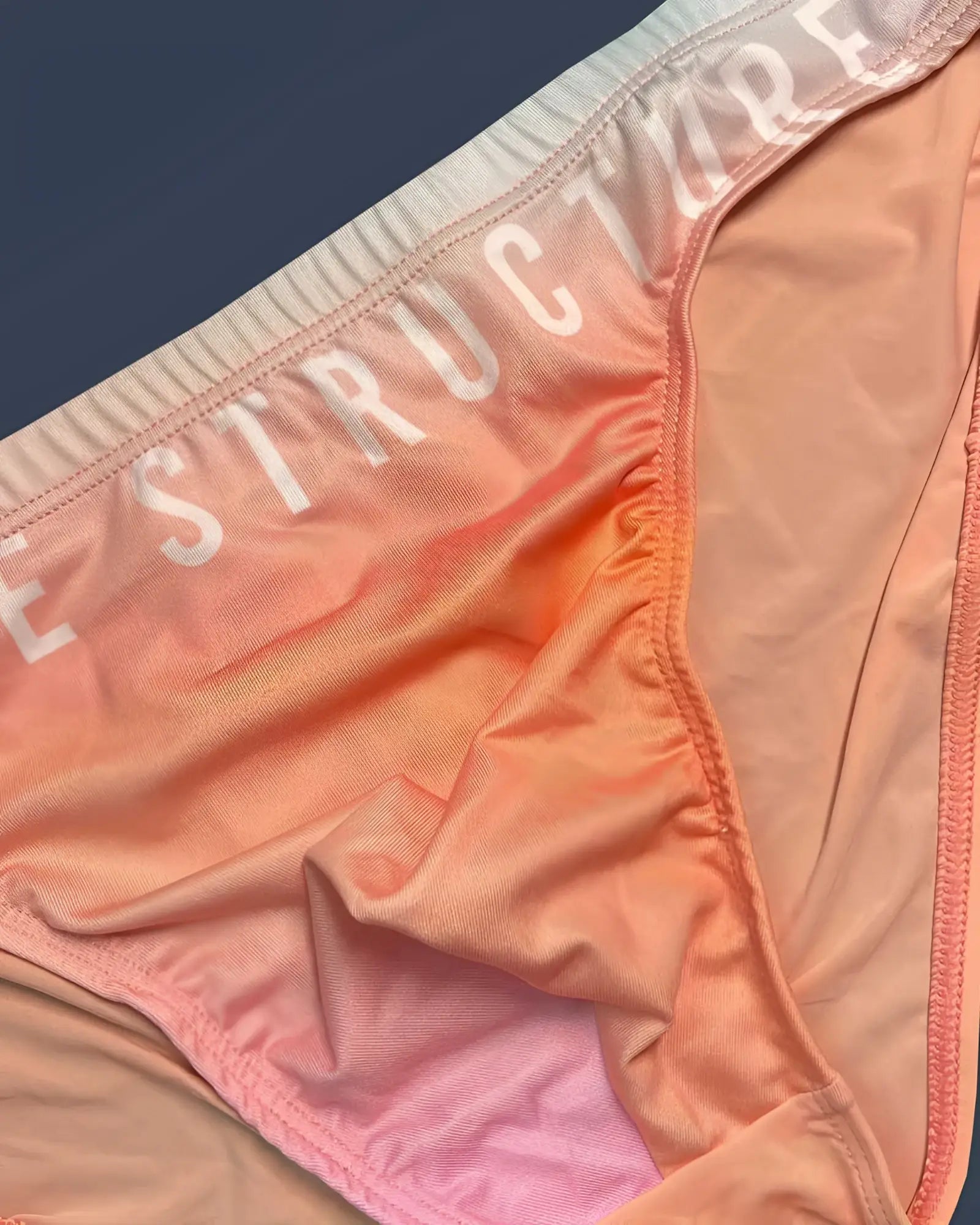 Peach and pink gradient fabric bikini bottom with structure printed white on waistband from private structure bare bikini in orange
