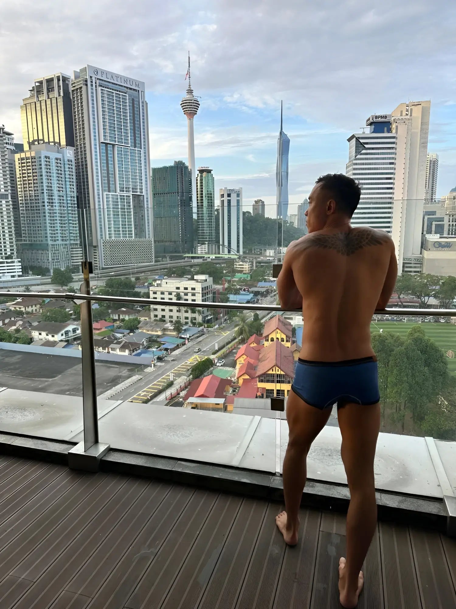 A man wearing dark blue denim Private Structure LCUT4359 barn boy low rise trunks stands on a wooden balcony