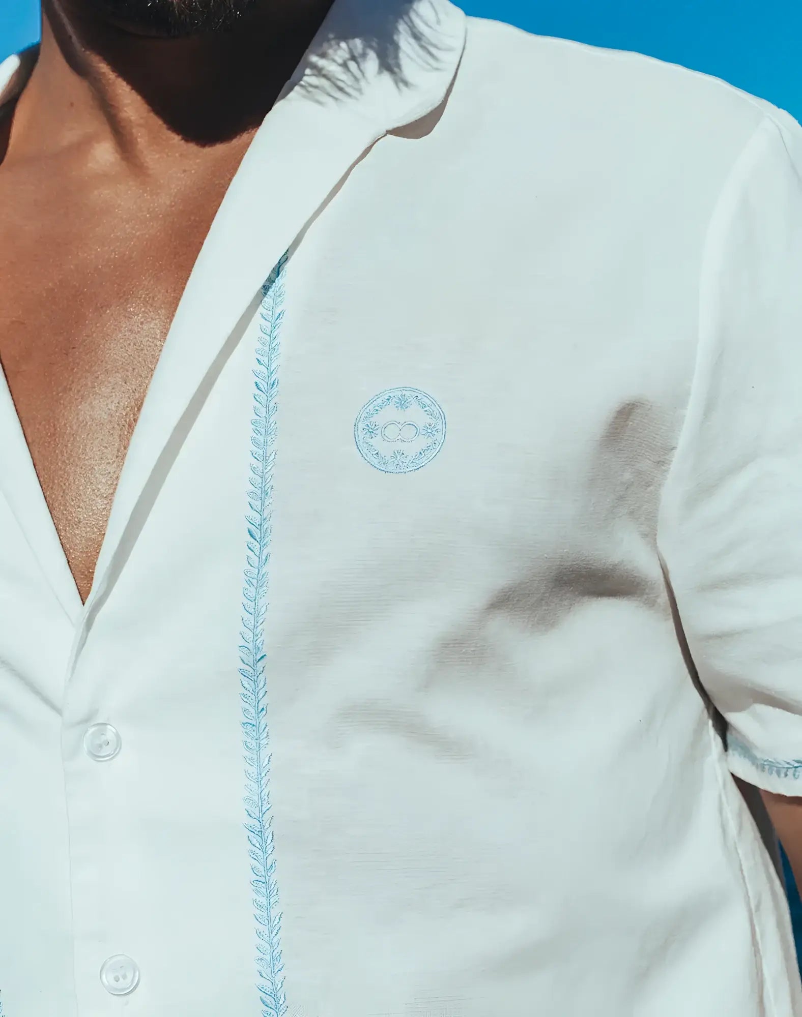 White santorini linen shirt with light blue embroidered detailing along the placket and matching logo on the chest