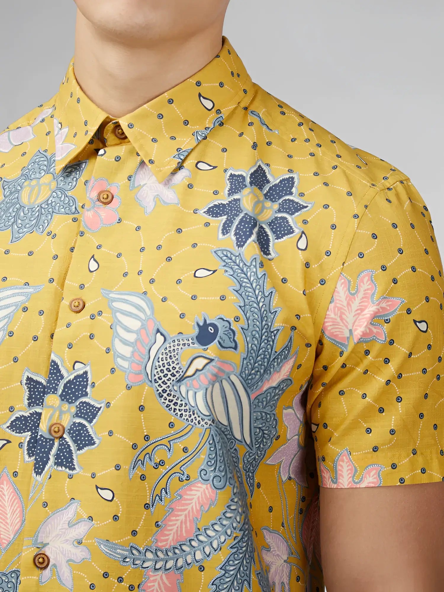 Yellow short-sleeved button-up shirt with intricate blue pink and white floral and bird patterns on textured fabric